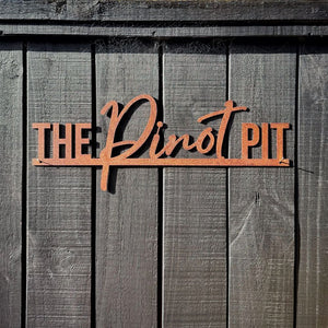 A rustic metal sign reading 'THE Pinot PIT' in corten steel, mounted on a dark wooden fence in Auckland.