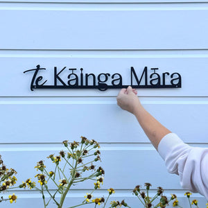 A black metal sign reading 'te Káinga Mára' mounted on a white horizontal-siding wall, with a person's hand adjusting it and small yellow-brown flowers in the foreground.