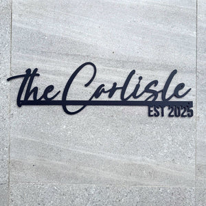 Black metal wall art featuring the script text 'the Carlisle' and the block text 'EST 2025' mounted on a light gray stone tile wall.