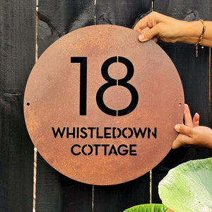 Corten steel custom round sign for traditional cottage in New Zealand on a black wooden fence. 