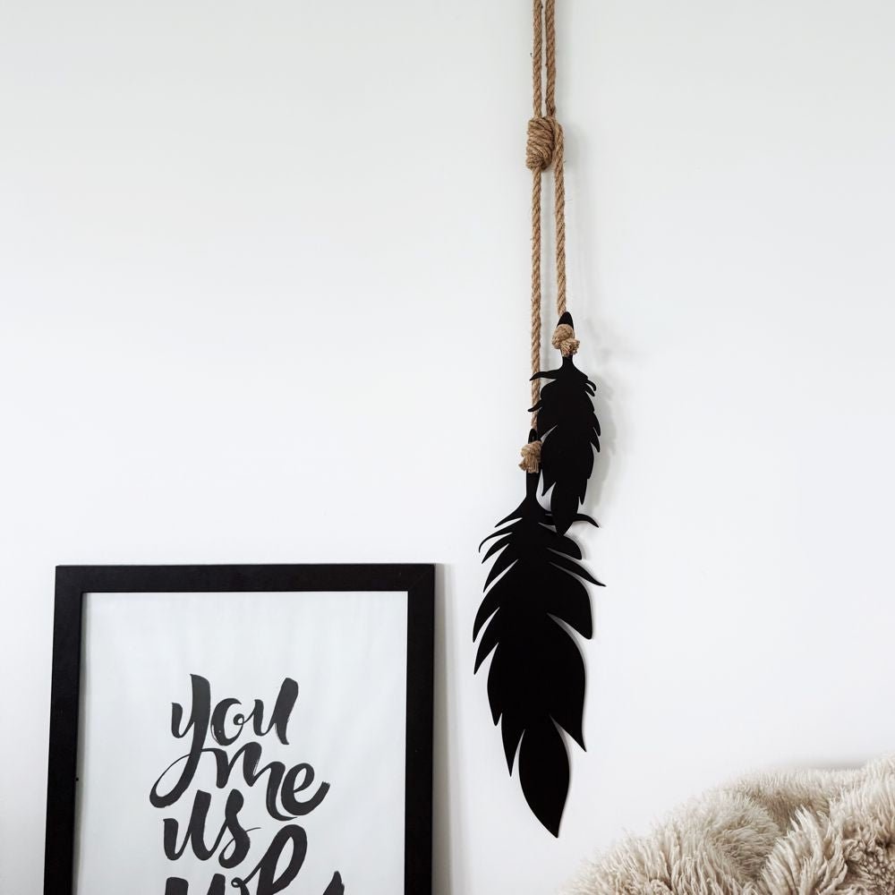 Black feather garden art hanging on a rope against a white wall with a framed picture below.