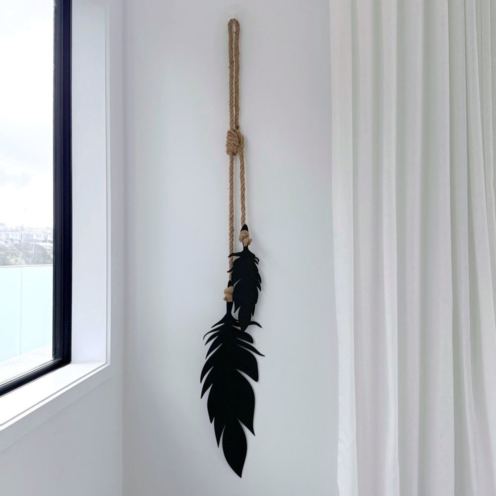 Black feather garden art hanging on a rope against a white wall with a framed picture below.