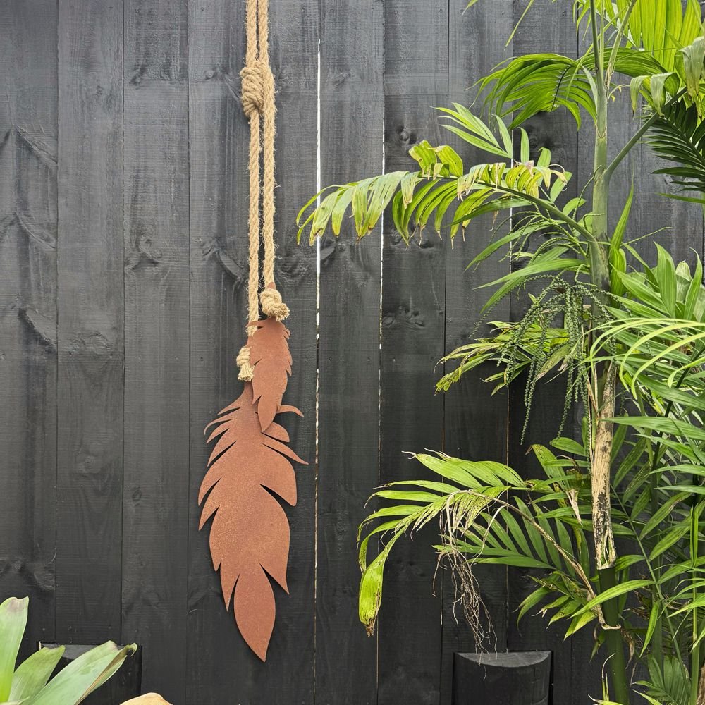 Large corten steel feather wall hanging displayed outdoors beside a pool with dog in New Zealand garden, premium metal art décor.
