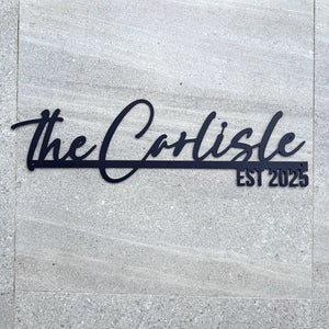 Black metal wall art featuring the script text 'the Carlisle' and the block text 'EST 2025' mounted on a light gray stone tile wall.