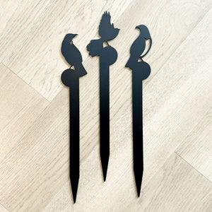 Three black metal garden stakes with bird designs on a light wood floor. LisaSarah