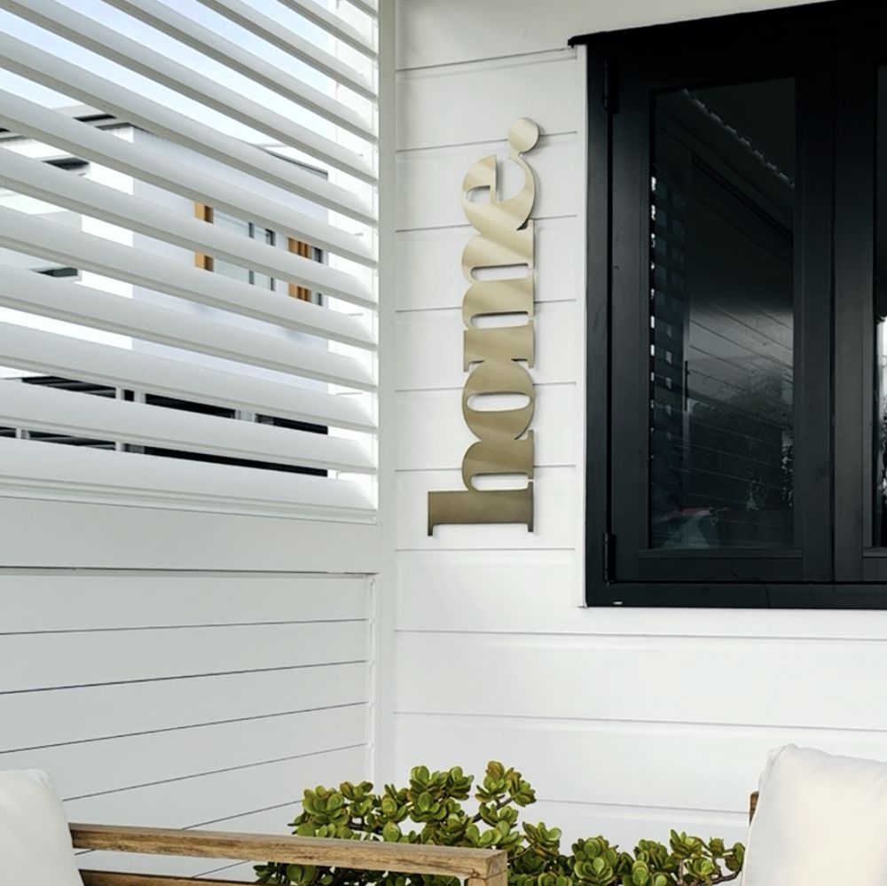 Decorative metal wall art spelling 'home' on a white wall next to a glass door with a plant. New Zealand outdoor living space. 