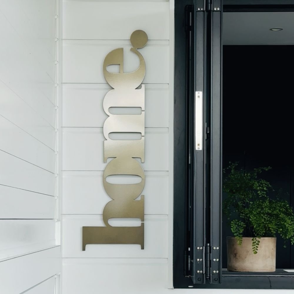 Decorative metal wall art spelling 'home' on a white wall next to a glass door with a plant. New Zealand outdoor living space. 