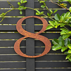 A large rustic metal number 8 with a weathered rust finish, mounted on a black wooden slatted fence and framed by green climbing vines.