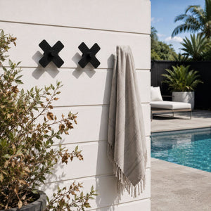 Large black steel cross wall hook by a pool holding a pool towel, NZ made