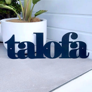 A black metal cutout sign spelling 'talofa' displayed on a light gray tiled surface in front of a white wall, with a white planter and green plant in the background.