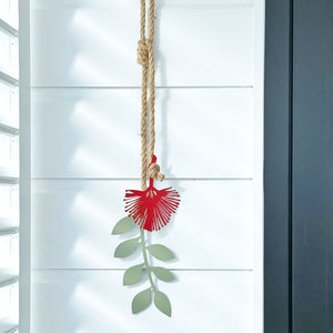 Red Pohutukawa flower garden ornament hanging from a rope on a white wall.