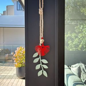 A decorative hanging pohutukawa ornament shaped like a red flower with pale green leaves, displayed on a dark window frame with an outdoor patio and garden visible in the background.
