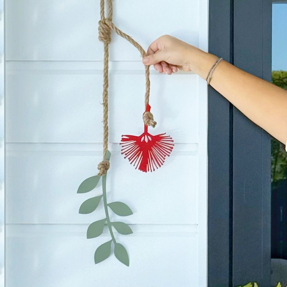 Decorative Pohutukawa flower with green leaves hanging on a rope against a dark wall with a pool and garden view.