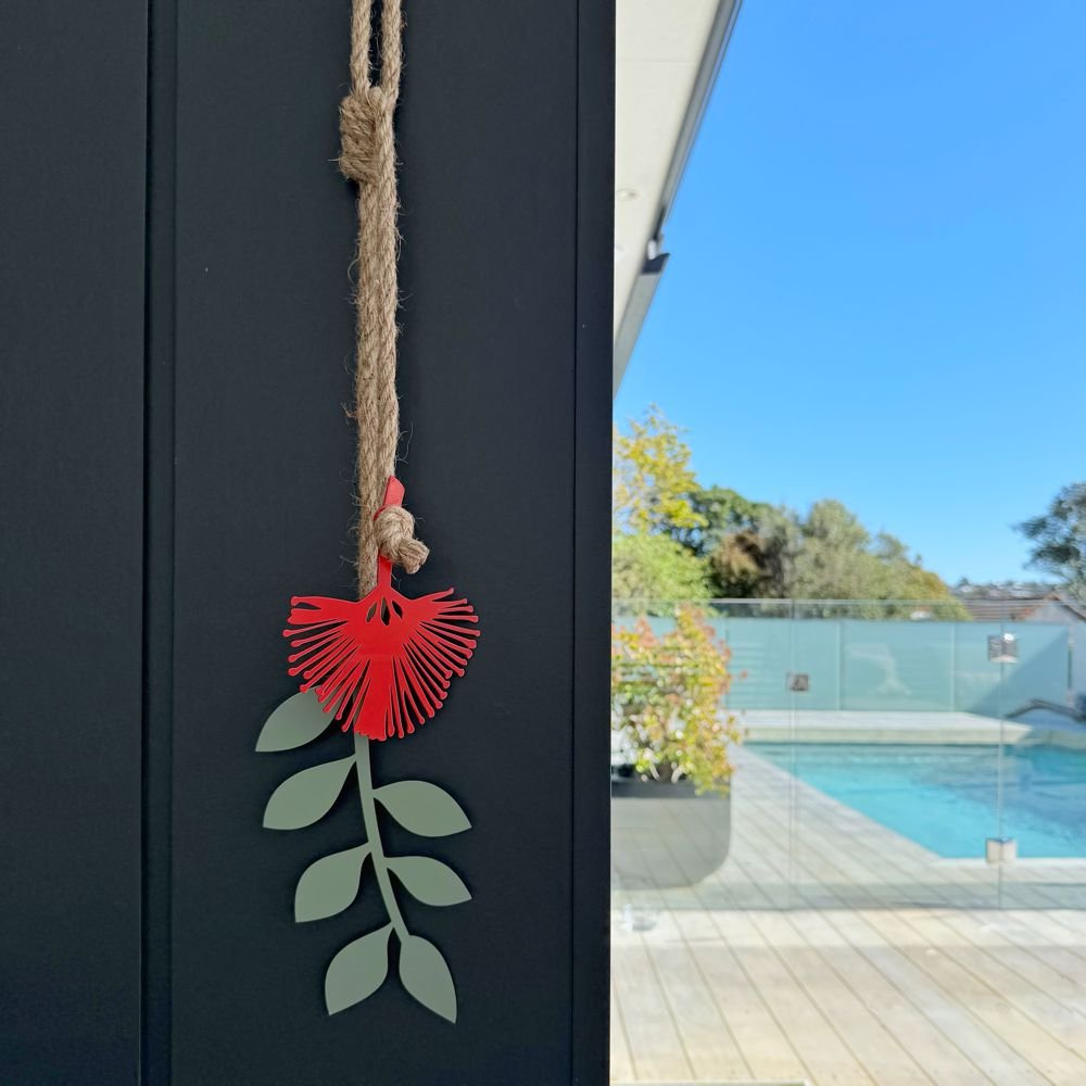 Decorative Pohutukawa flower with green leaves hanging on a rope against a dark wall with a pool and garden view.