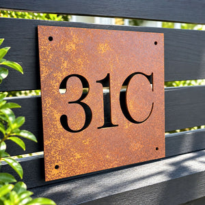 A square rusted metal house number sign reading '31C' mounted on a dark wooden slat fence, with green foliage visible on the left.