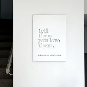 A minimalist typographic wall art print hanging on a white wall, featuring the phrase 'tell them you love them.' in large gray letters and a smaller line below reading 'tell them a lot, make it weird.