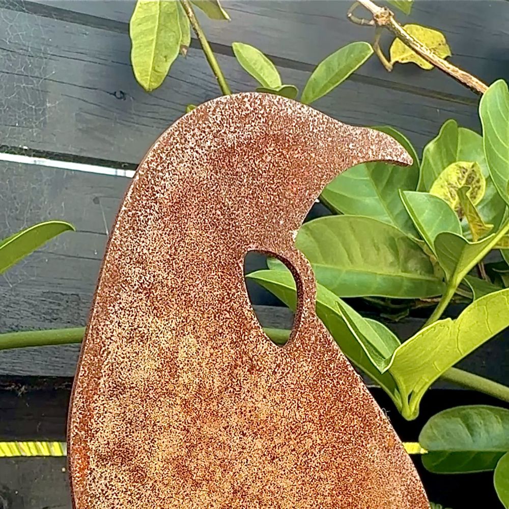 A rusted tui metal garden stake, displayed outdoors against a dark wooden fence with green plants.