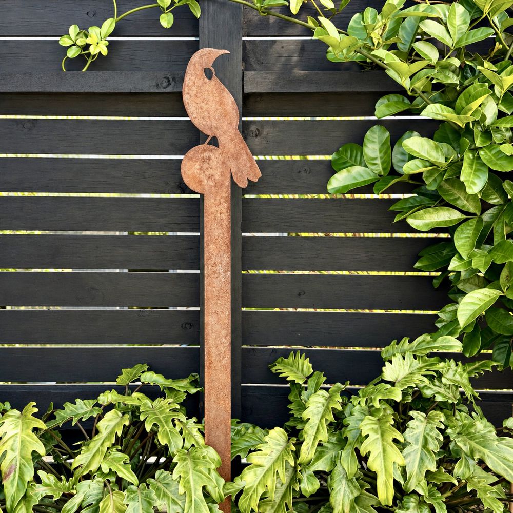 A rusted tui metal garden stake, displayed outdoors against a dark wooden fence with green plants.