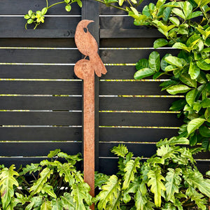 A rusted tui metal garden stake, displayed outdoors against a dark wooden fence with green plants.