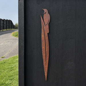 Corten steel Tūī on flax garden stake mounted on a black fence as wall art in a New Zealand backyard